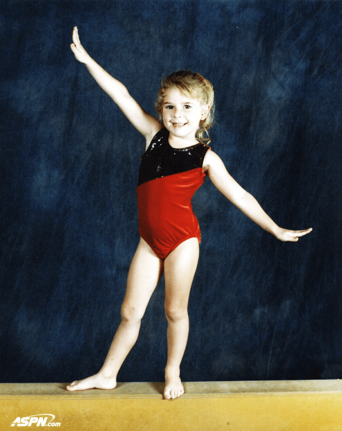 A young girl in a red and black leotard is standing on one foot.