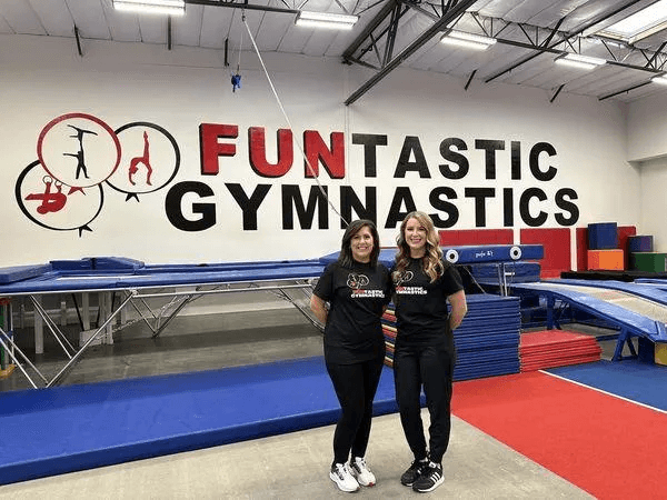 Two women standing in front of a gym with the words " funtastic gymnastics ".